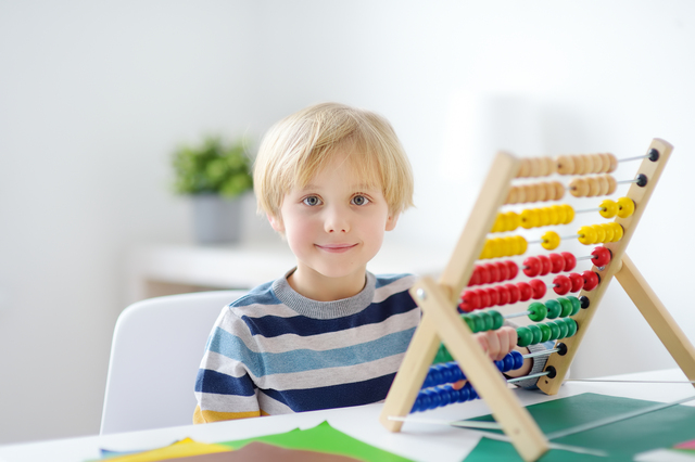 外国人の小学生の男の子が大きなそろばんを机において、カメラに向かって微笑んでいる。