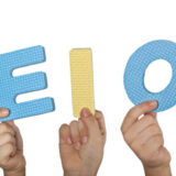 アルファベットの、A、E、I、O、Uの文字を手に持っている。