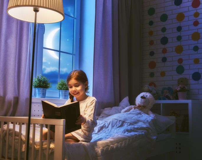 6歳くらいの女の子がベッドの上に座って月明かりのもとで本を読んでいる。