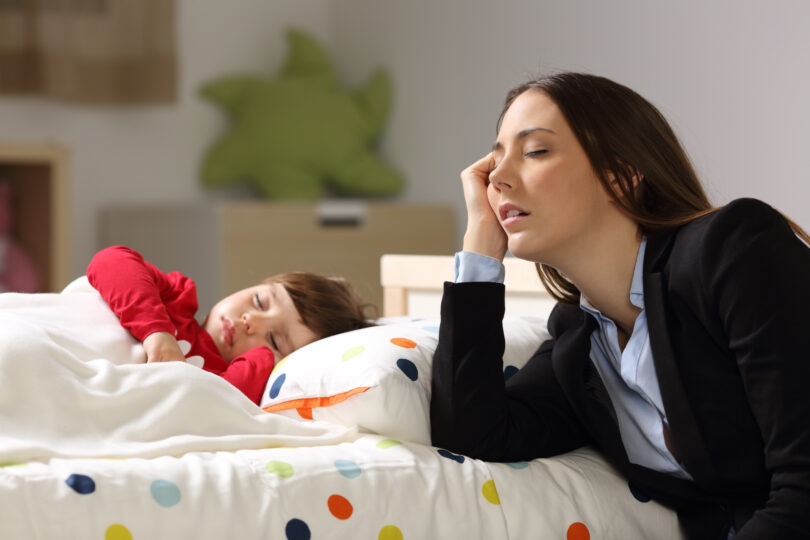 仕事で疲れて帰ってきたスーツ姿の母親の横で女の子がスヤスヤと眠っている。