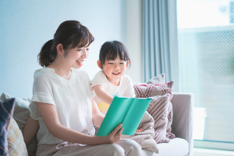 アジア系の母親と子どもがソファで絵本を読んでいる。